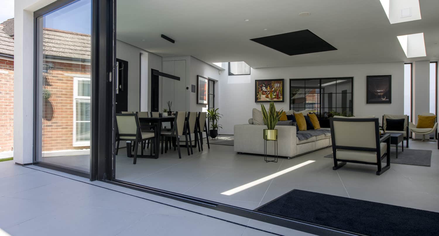 Modern open-plan living and dining room with expansive bifold doors opening to a patio, featuring neutral-toned furniture, plants, wall art, and abundant natural light.