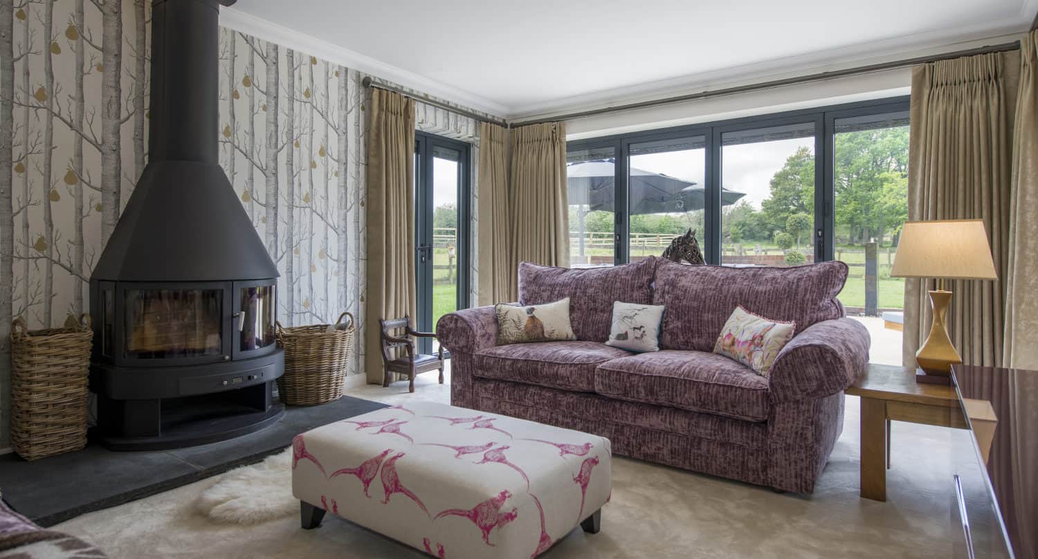 A cozy living room with a purple sofa, floral ottoman, modern black wood-burning stove, patterned wallpaper, large windows, and bifold doors opening to views of a green lawn and trees outside.