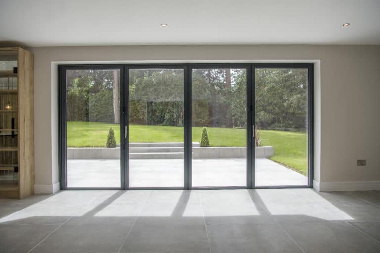 Large floor-to-ceiling bifold doors open to a patio and a green garden with trees and manicured grass, letting natural light flood the modern, tiled interior space.