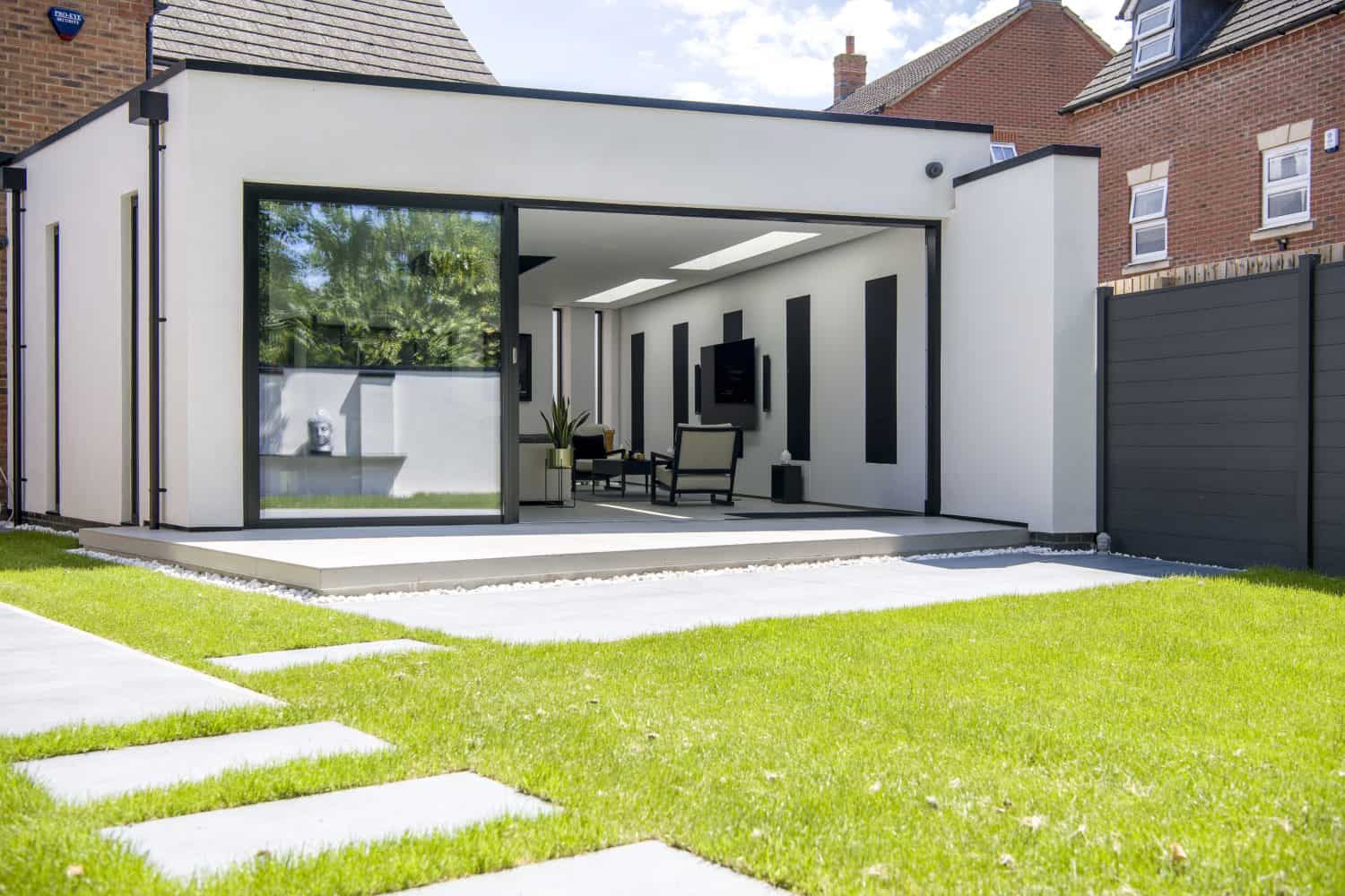 Modern home extension with bifold doors opening to a patio and green lawn, featuring minimalistic furniture inside and surrounded by a wooden fence and neighboring brick houses.