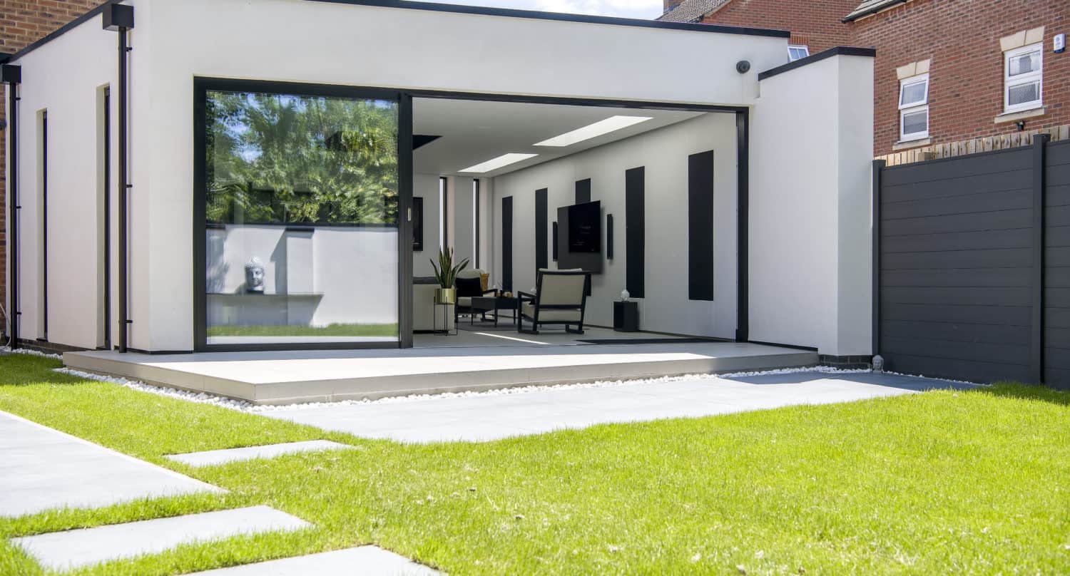 Modern home extension with bifold doors opening to a patio and green lawn, featuring minimalistic furniture inside and surrounded by a wooden fence and neighboring brick houses.