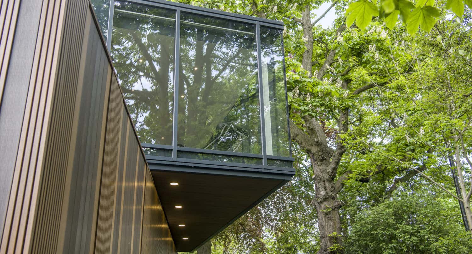 Modern building with a cantilevered glass room and bifold doors, surrounded by lush green trees reflecting in the glass. The lower wood-paneled wall features recessed lights. The scene appears bright and natural.