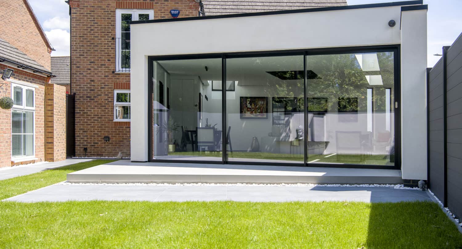 Modern home extension featuring bespoke glass solutions with expansive floor-to-ceiling sliding doors, a white exterior, flat roof, and small raised patio—perfectly complementing the adjacent brick house and lush green lawn.
