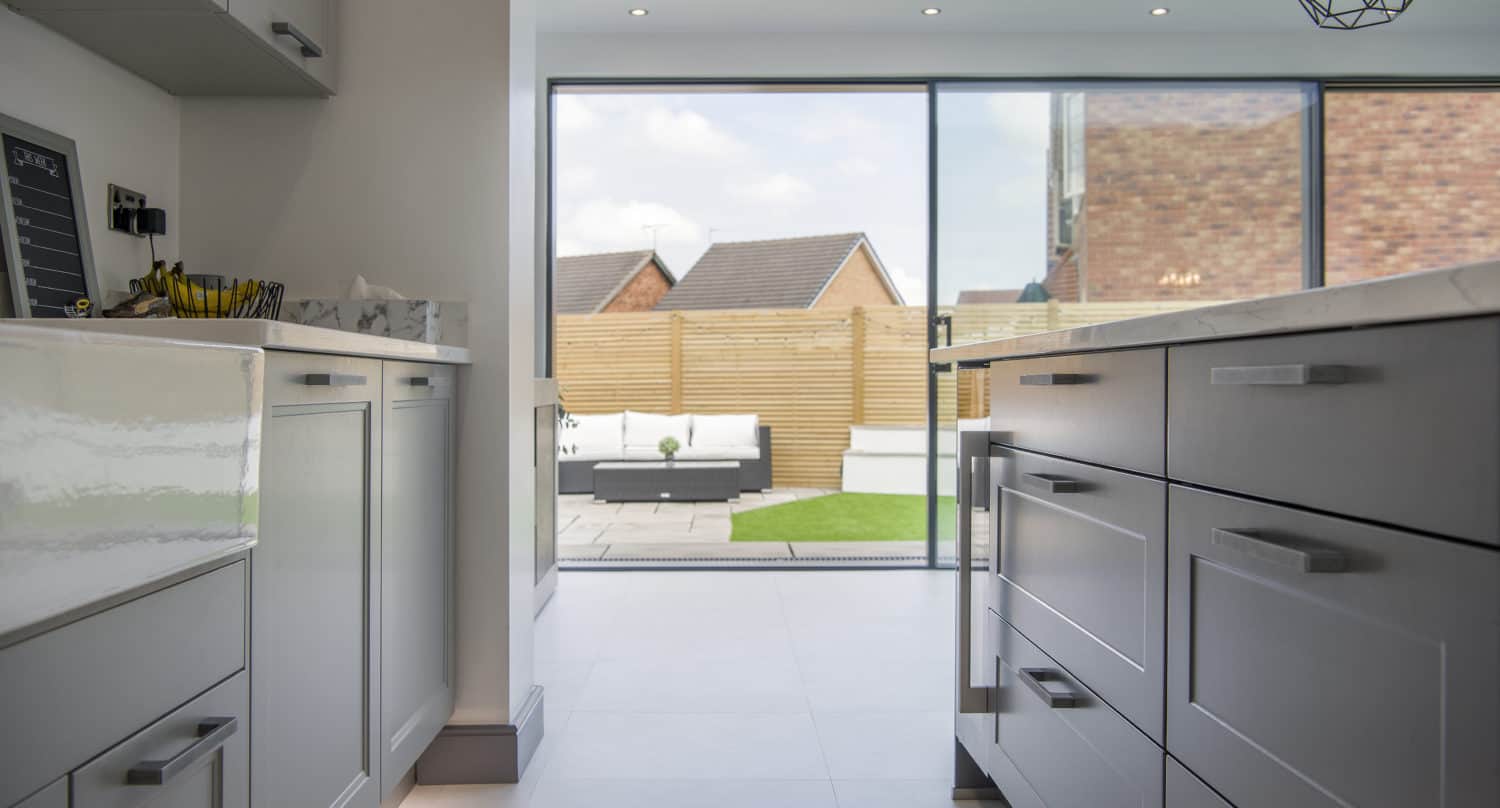 Modern kitchen with light gray cabinets and marble countertops, opening to a patio with outdoor seating and a fenced yard, visible through expansive bifold doors. Natural light fills the space.