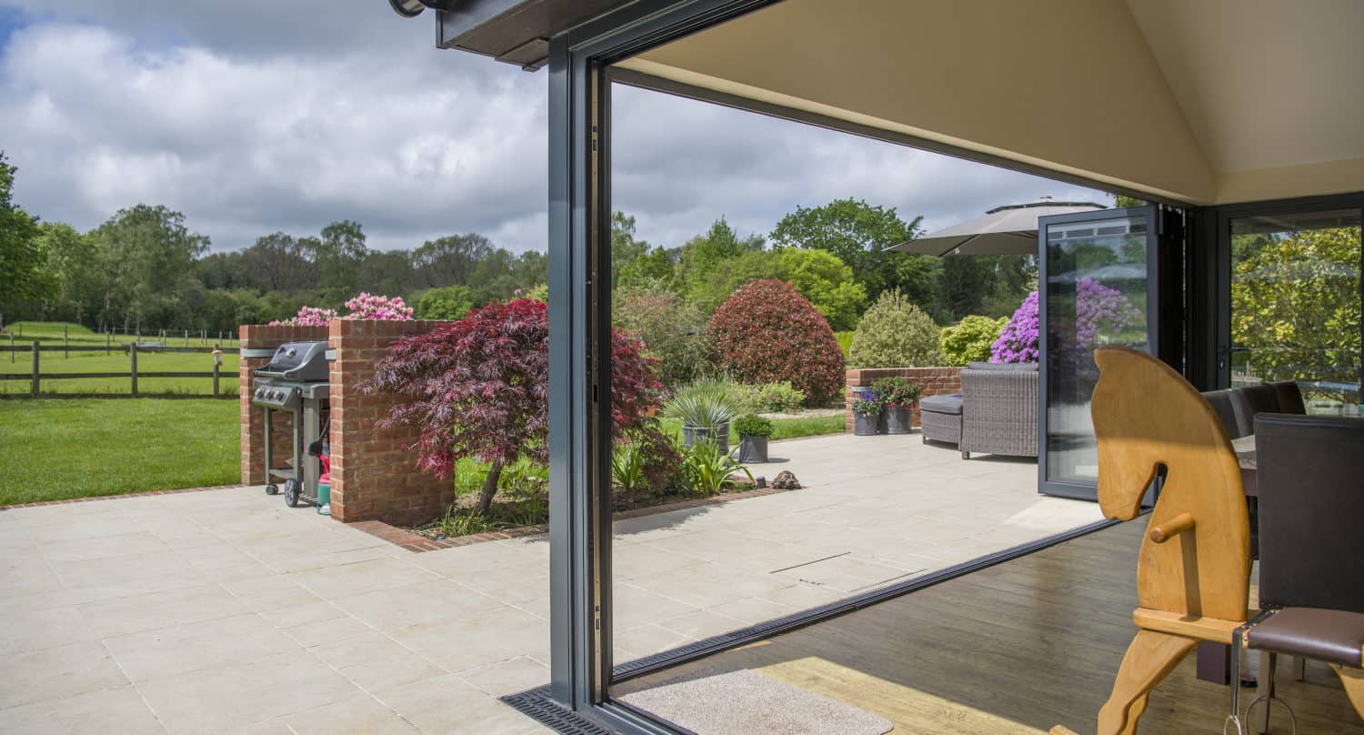 View from inside a house through expansive bifold doors to a patio with outdoor seating, a barbecue grill, and a garden filled with colorful shrubs and green trees under a partly cloudy sky.