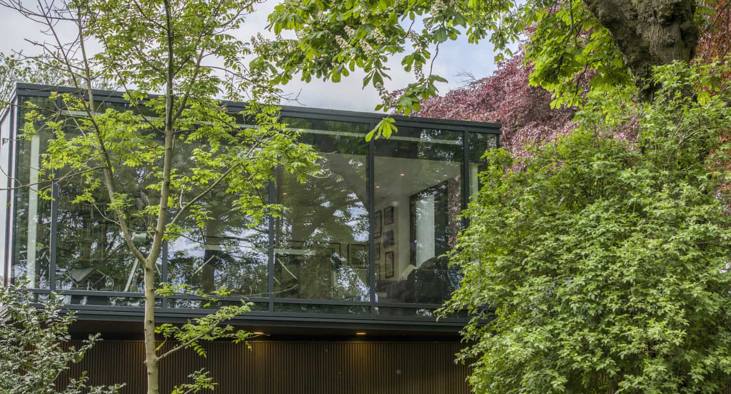 Modern glass-walled house surrounded by lush green trees and foliage, with bespoke glass solutions like large windows and bifold doors reflecting the sky. The home blends seamlessly into its natural environment.