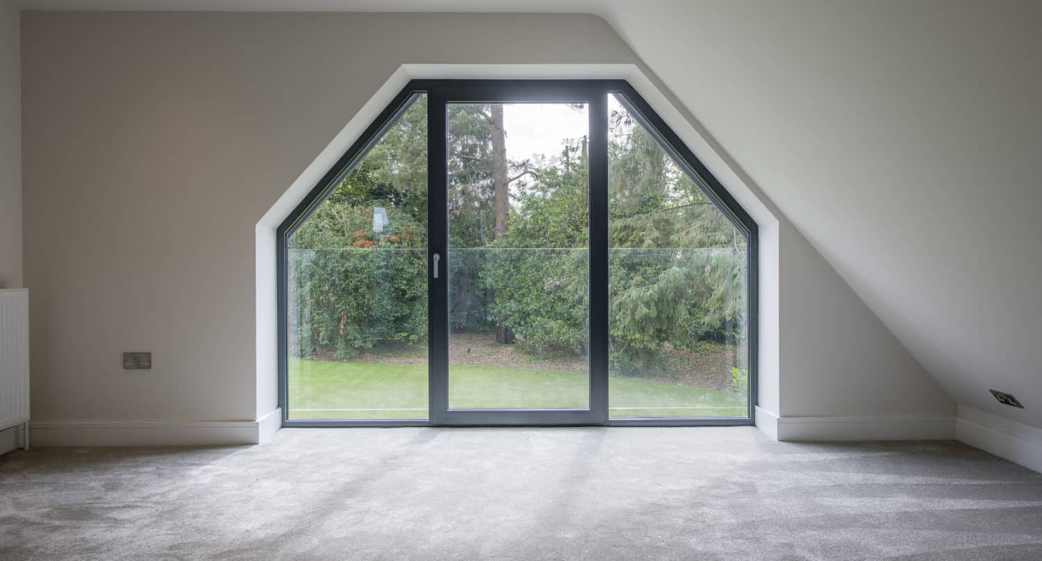 A bright room with grey carpet, featuring large bifold doors with side windows opening to a garden view, framed by white walls and a sloped ceiling.