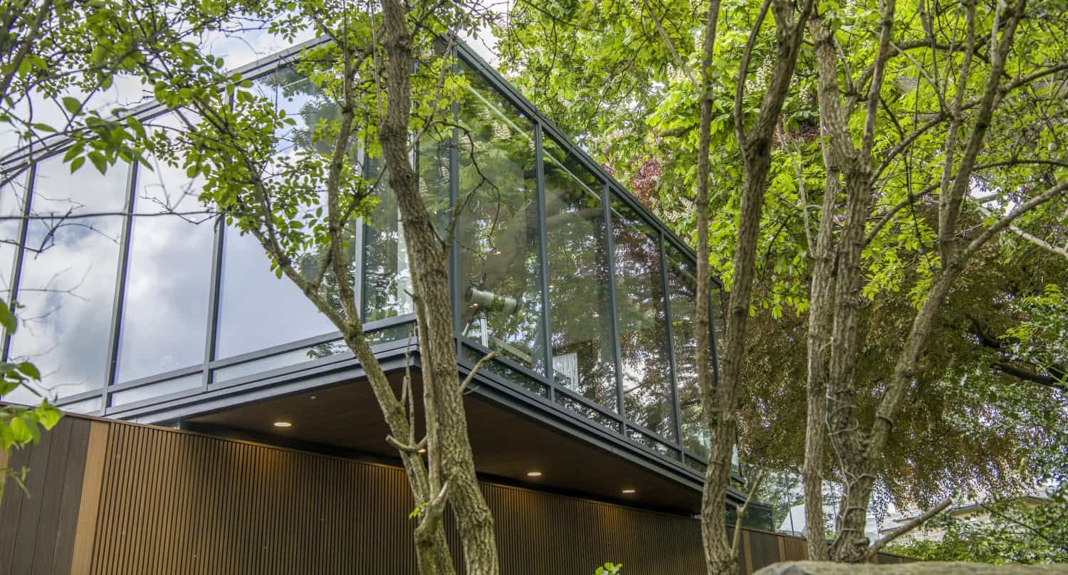 Modern glass building with bespoke glass solutions, surrounded by lush green trees, reflecting the sky and foliage. Features a wooden lower facade, overhanging upper section, and elegant sliding doors that invite nature inside.