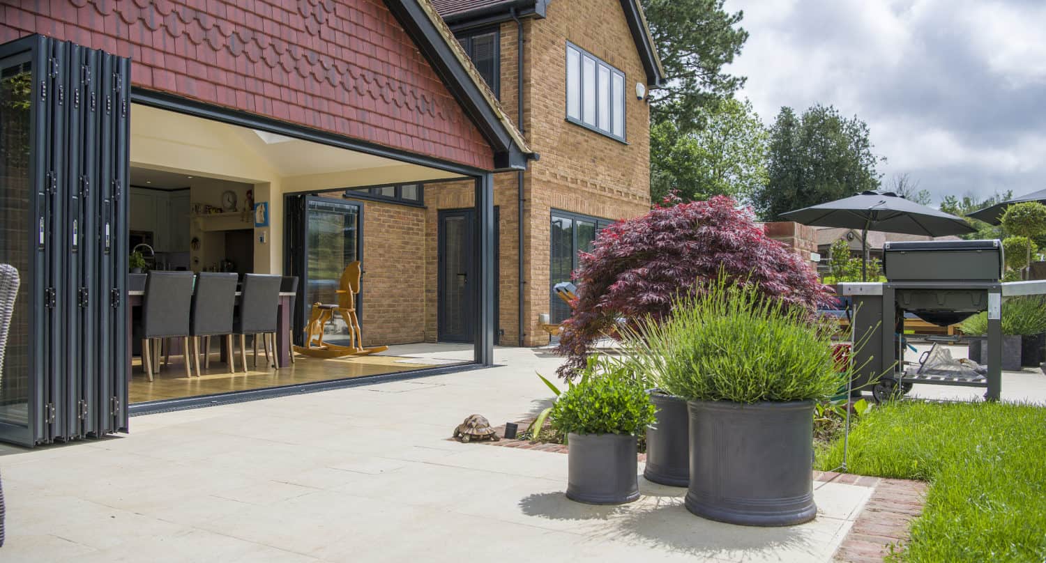 A modern house with bespoke glass solutions, including large glass folding doors opening to a sunny patio with potted plants, outdoor dining furniture, and a garden featuring green grass and trees in the background.
