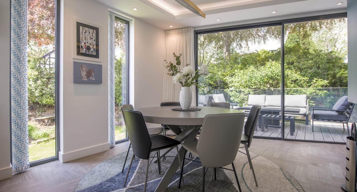 A modern dining room with a round table, six chairs, a patterned rug, large windows, and bespoke glass sliding doors opening to a patio with outdoor seating. A vase of flowers sits on the table, and greenery is visible outside.