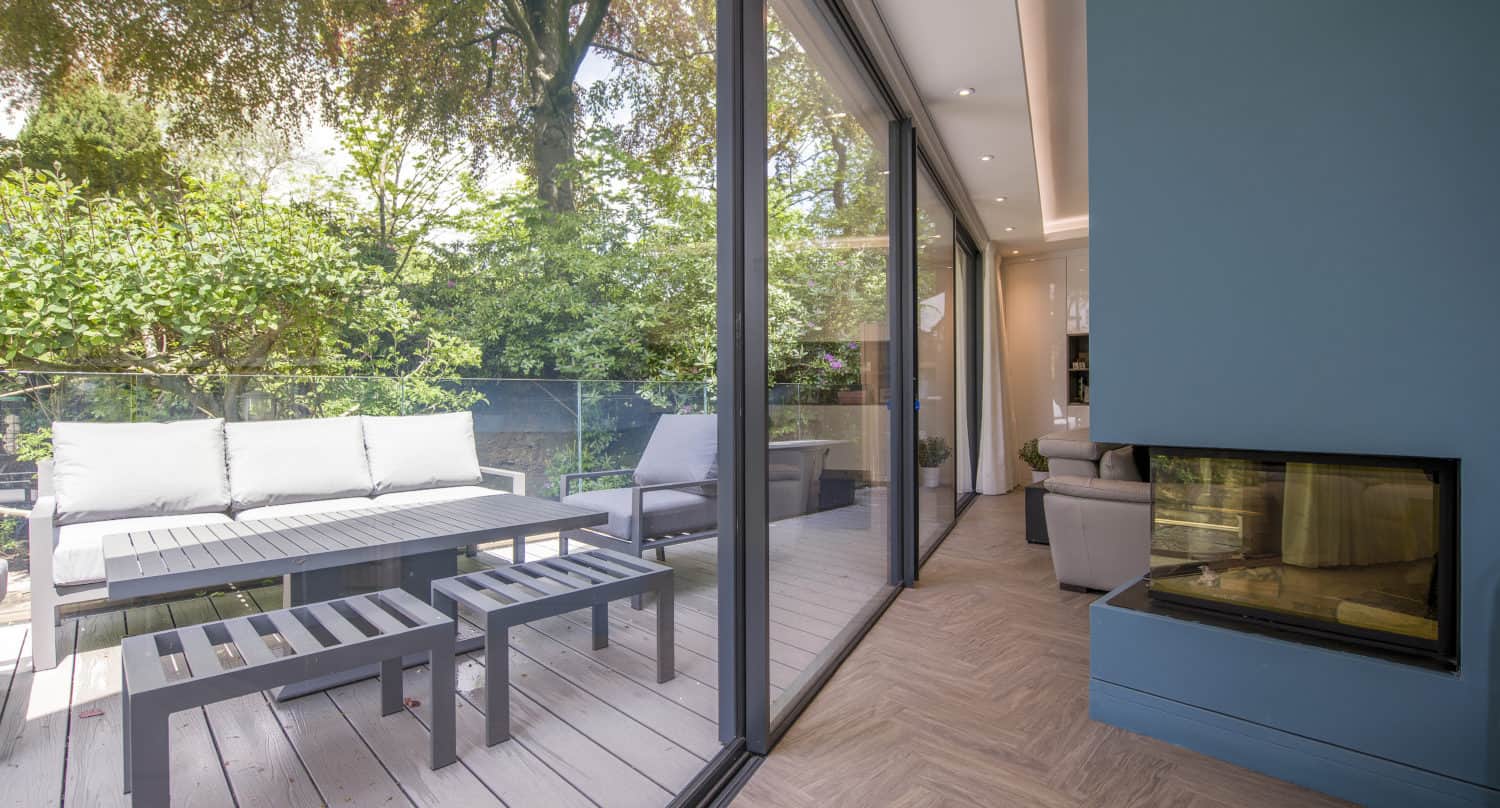 A modern living space with sliding doors opens onto a wooden deck with gray outdoor furniture, surrounded by lush green trees and plants. A corner fireplace is visible inside the blue-walled room.