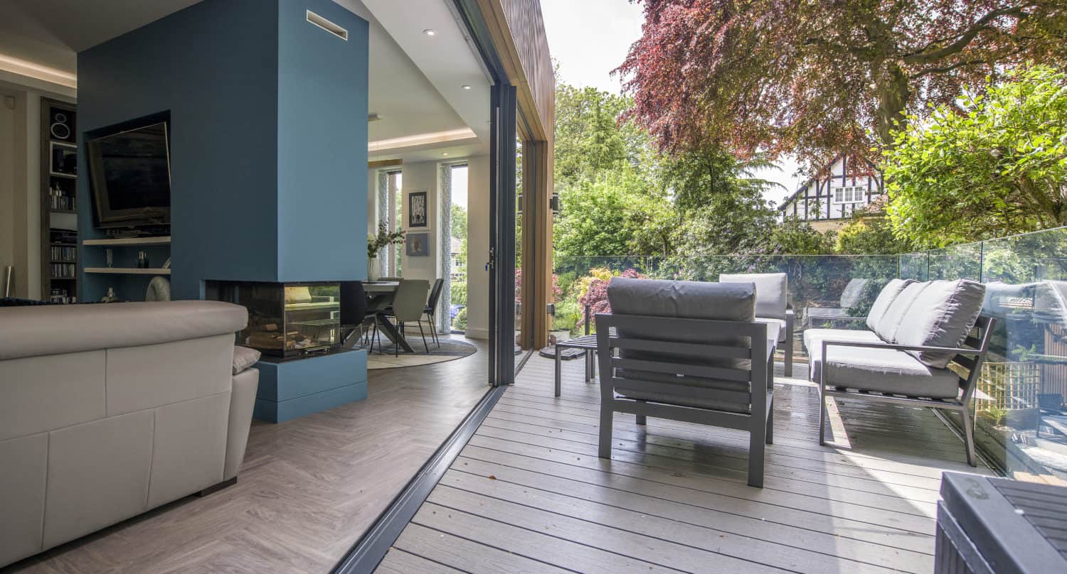A modern living room with light-colored furniture opens onto a spacious patio with seating. Bifold doors connect indoor and outdoor areas, overlooking a lush garden filled with trees and greenery.