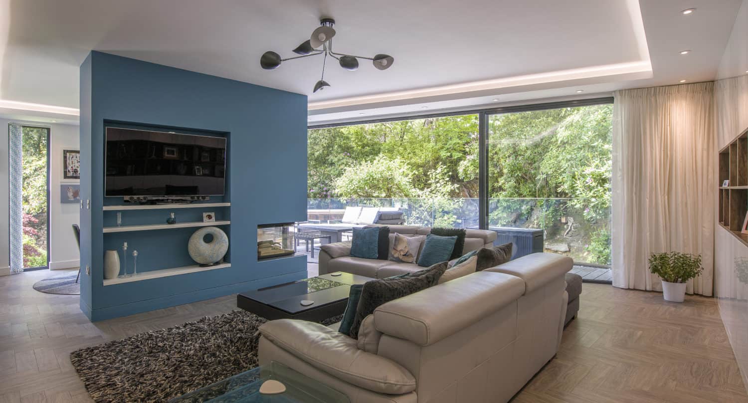 Modern living room with light leather sofas, a blue accent wall with a mounted TV, shaggy rug, glass coffee table, and large bifold doors offering seamless views of the green, tree-filled outdoor space.