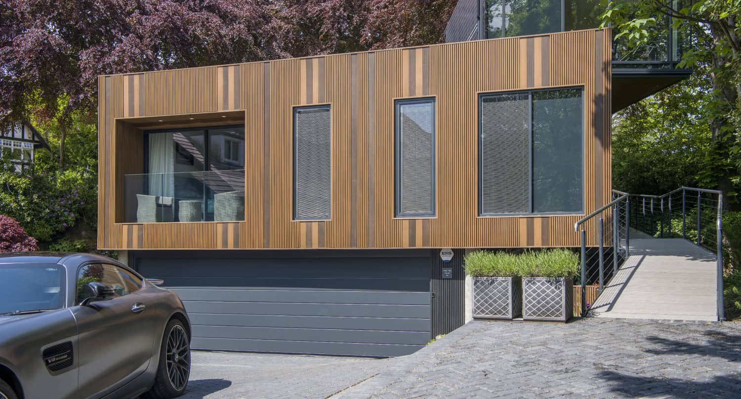 A modern house with a wooden exterior and large windows sits above a gray garage. Sliding doors open to the outdoors, while a black car is parked in the driveway, with trees and greenery surrounding the home.
