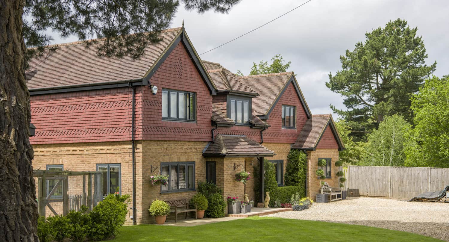 A large brick and red-tiled house with multiple gables, black-framed windows, bifold doors, potted plants, hanging flower baskets, and a well-kept lawn, surrounded by trees and a gravel driveway under a partly cloudy sky.