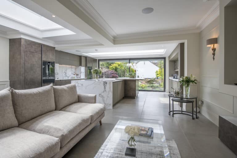 Modern open-plan living space with a light beige sofa, glass coffee table, and a stylish kitchen featuring marble countertops. Large bespoke glass solutions, including sliding front doors, open to a lush garden and flood the area with natural light.