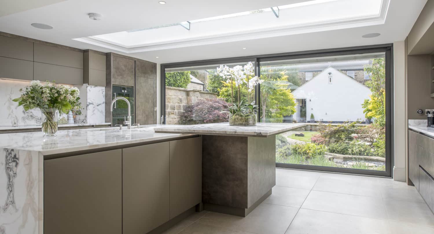 Modern kitchen with marble countertops, large island, and floor-to-ceiling bifold doors opening to a landscaped garden with a white house visible outside. Natural light floods the space through a skylight.