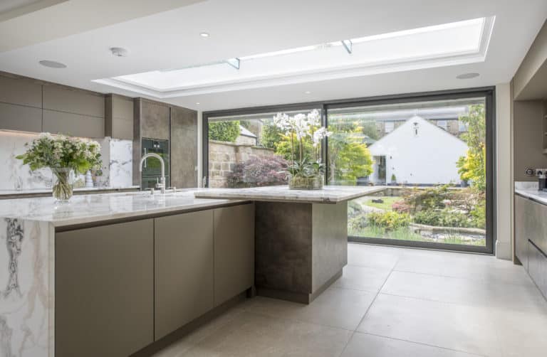 Modern kitchen with marble countertops, large island, and floor-to-ceiling bifold doors opening to a landscaped garden with a white house visible outside. Natural light floods the space through a skylight.