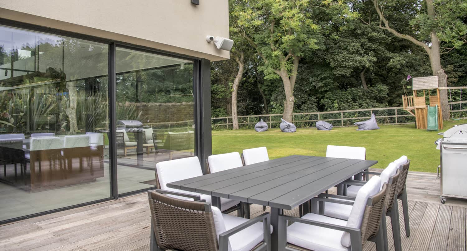 Outdoor patio with a dark dining table and eight cushioned chairs on a wooden deck, next to a modern house featuring bespoke glass solutions and large glass doors. The garden has green grass, trees, bean bags, and a children’s playhouse.