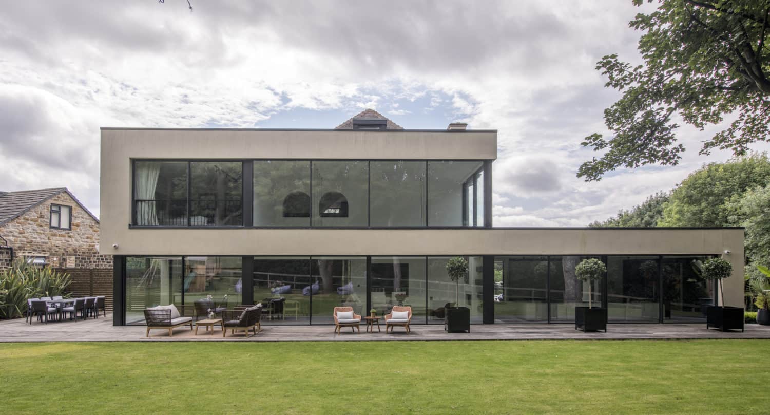 Modern two-story house with bespoke glass solutions, featuring large windows, a spacious wooden patio with outdoor seating, and a well-kept lawn in the foreground. Trees and greenery surround the contemporary building.