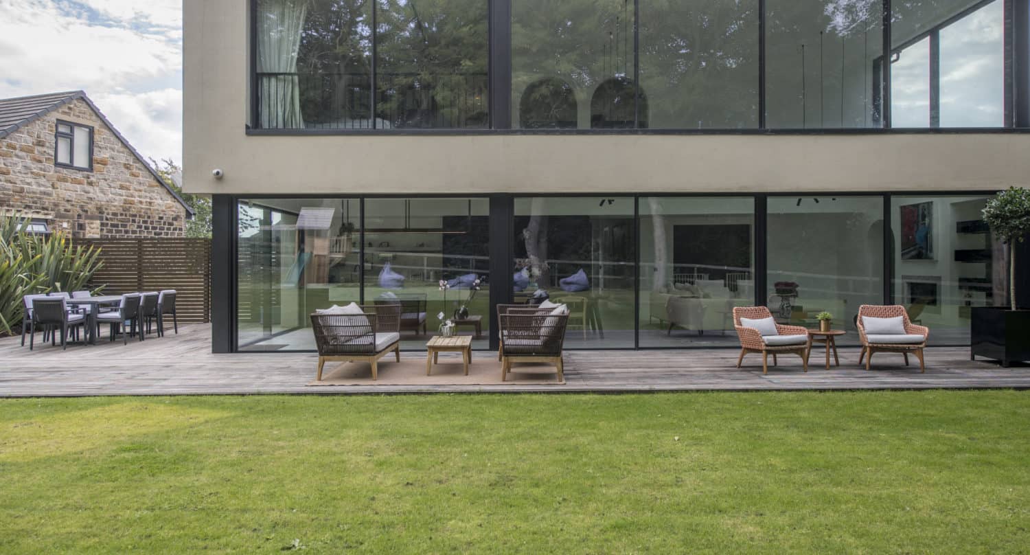 Modern house with large glass windows and bespoke glass solutions, plus a wooden patio featuring outdoor seating. There are armchairs and a table on the deck, a dining table to the side, and a well-maintained lawn in the foreground.