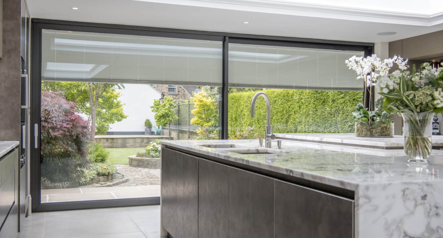 Modern kitchen with a marble island, built-in sink, and bespoke sliding doors leading to a landscaped garden. Natural light streams in through a skylight, highlighting potted orchids on the countertop.