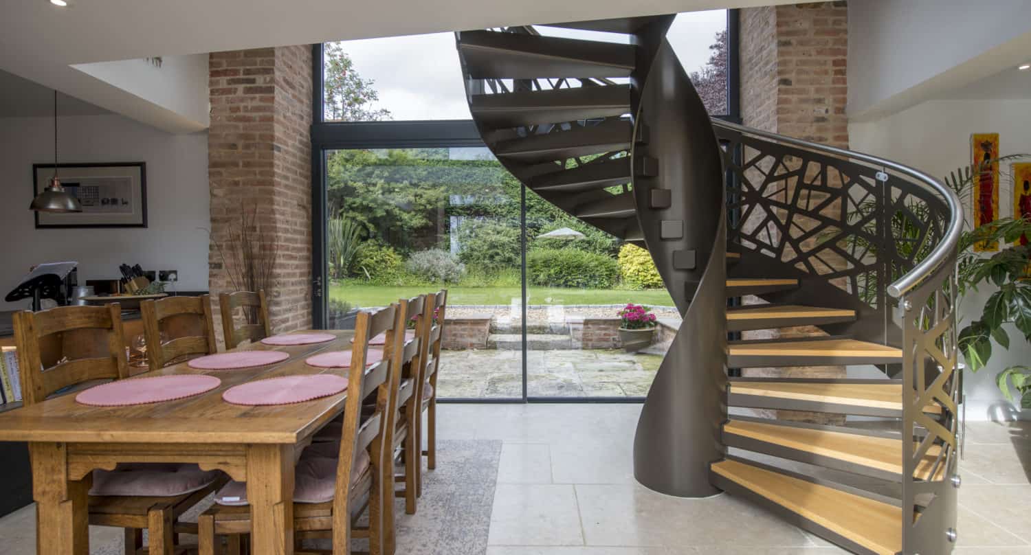 A modern dining area with a wooden table and chairs sits beside a stylish spiral staircase with geometric railings, bespoke glass solutions for the large doors, and a view of the green garden outside.