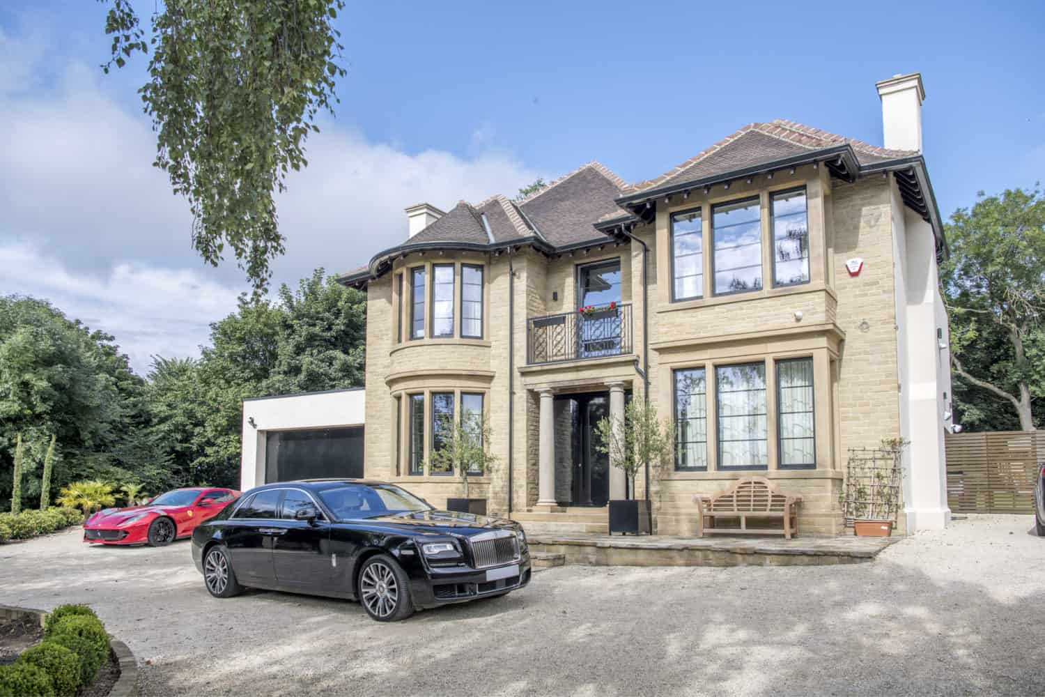 A large, modern two-story house with tan brick walls, large windows, and sleek sliding doors. A black luxury car and a red sports car are parked in the driveway, with trees and greenery surrounding the property.
