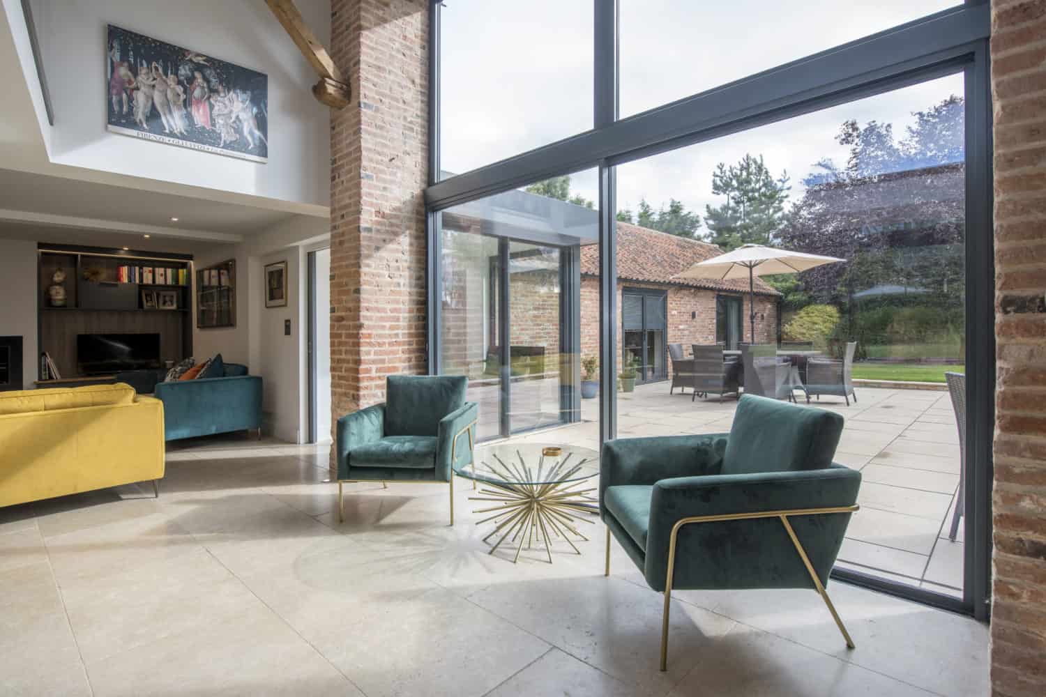 Modern living room with large windows, green velvet chairs, a yellow sofa, and a round glass coffee table. Sliding doors open to a patio with outdoor furniture and garden views. Exposed brick and artwork adorn the interior.