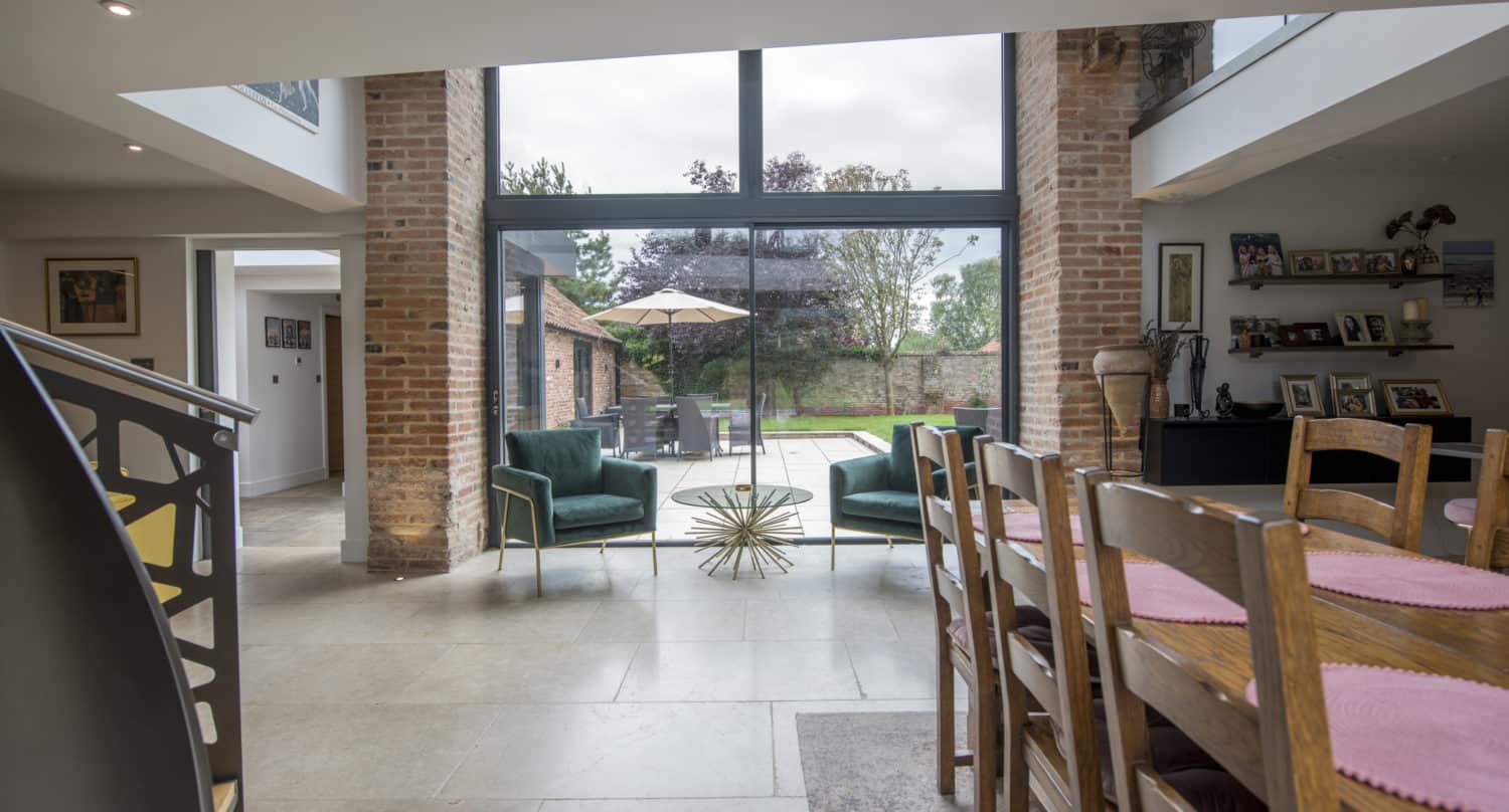 Modern dining and living area with large bespoke glass sliding doors opening to a patio. Wooden dining table with chairs, green armchairs by a gold coffee table, brick walls, and outdoor seating visible through the windows.