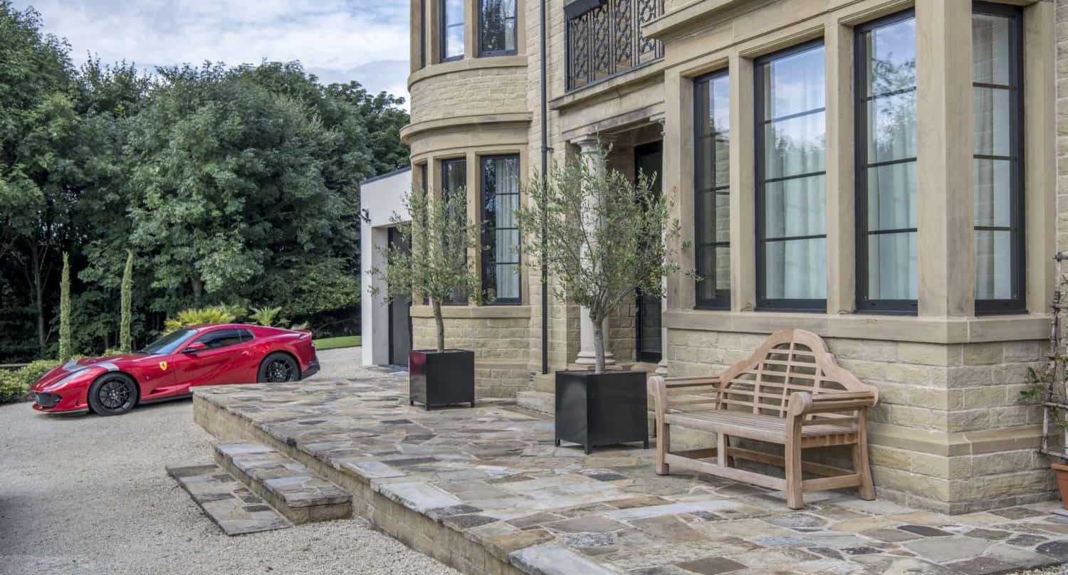 A red sports car is parked on a gravel driveway next to a large stone house with tall windows, bifold doors, potted plants, and a wooden bench near the entrance. Trees and greenery are in the background.