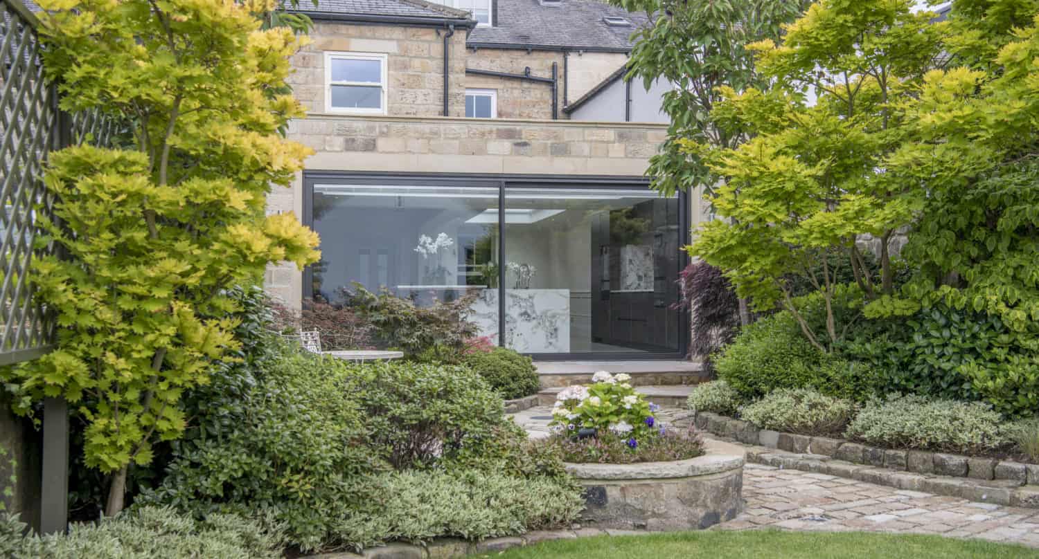 A lush garden with green shrubs, curved stone paths, and flowering plants leads to a modern house extension featuring bespoke glass solutions, large sliding doors, and a view into a bright, contemporary interior.