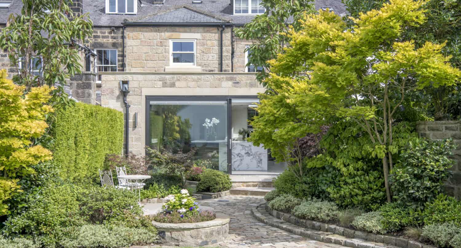 A stone house with large bifold doors opens to a lush garden, featuring a winding stone path, green lawn, vibrant shrubs, trees, and a small circular patio with white garden chairs and a table.