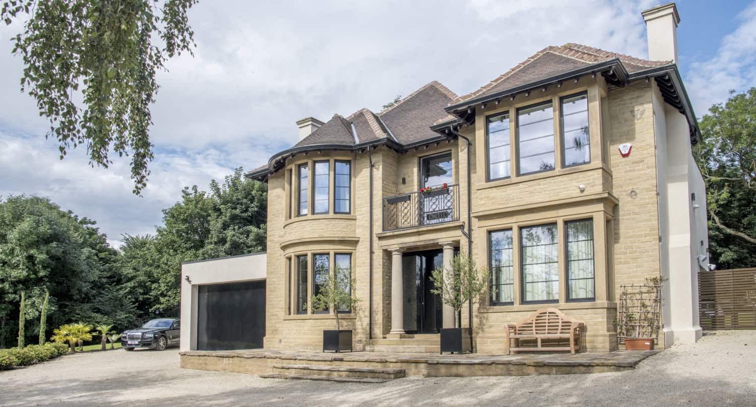 Large two-story beige stone house with multiple big windows, bifold doors, a double garage, and a curved driveway. Trees and shrubs surround the property, with a bench by the entrance and a car parked on the left side.