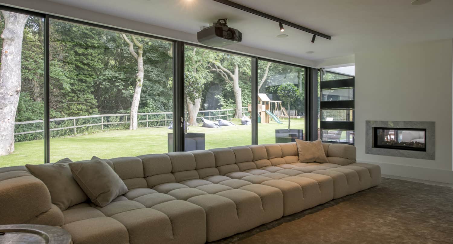 A modern living room with a long beige sectional sofa, large sliding doors overlooking a green backyard with trees and a playset, and a built-in fireplace on the right wall.