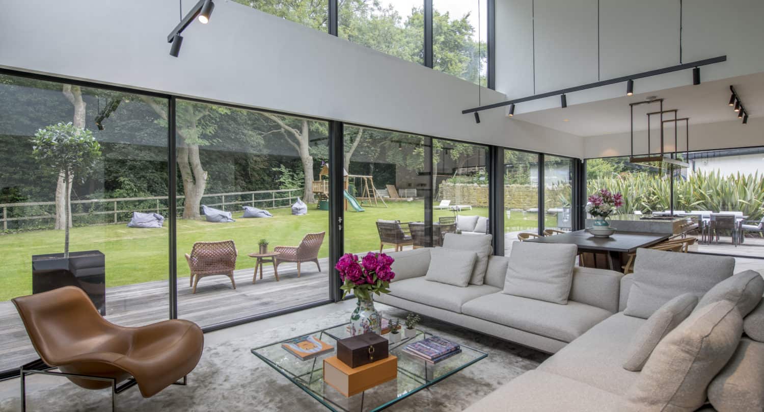 A modern living room with large bespoke glass solutions for floor-to-ceiling windows overlooking a green yard. The space features a light gray sectional sofa, a brown lounge chair, and a glass coffee table with pink flowers and books.