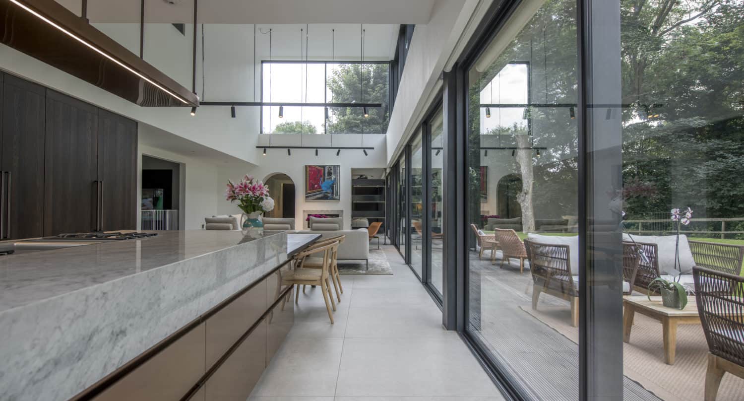 Modern kitchen with a marble island and wooden cabinets, leading to a bright living area. Large bespoke glass solutions, including sliding doors, open to a patio with outdoor seating and views of trees and greenery.