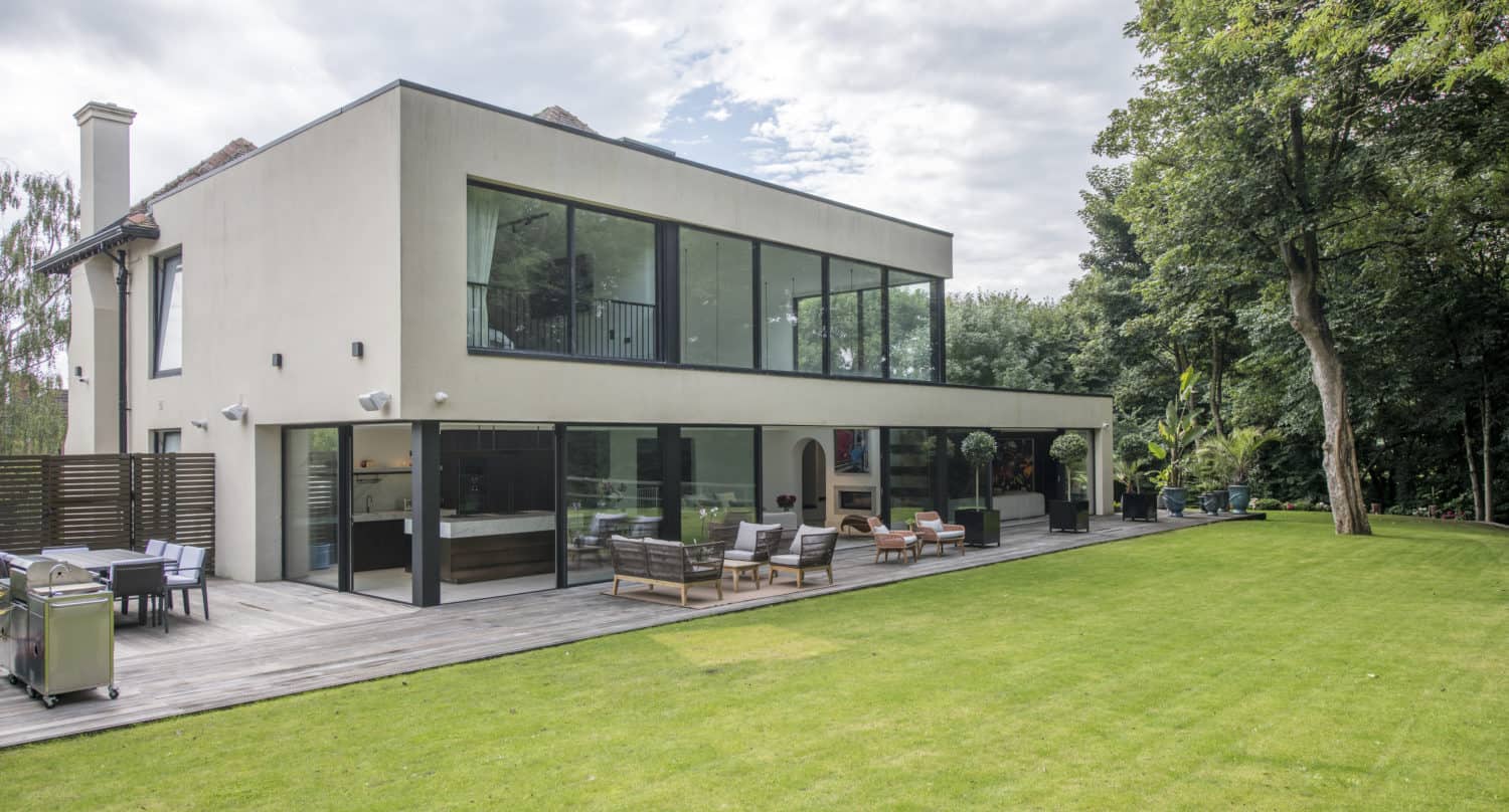 A modern two-story house with large windows, sliding doors opening to an outdoor seating area on a wooden deck, and a spacious, well-maintained lawn bordered by trees under a partly cloudy sky.