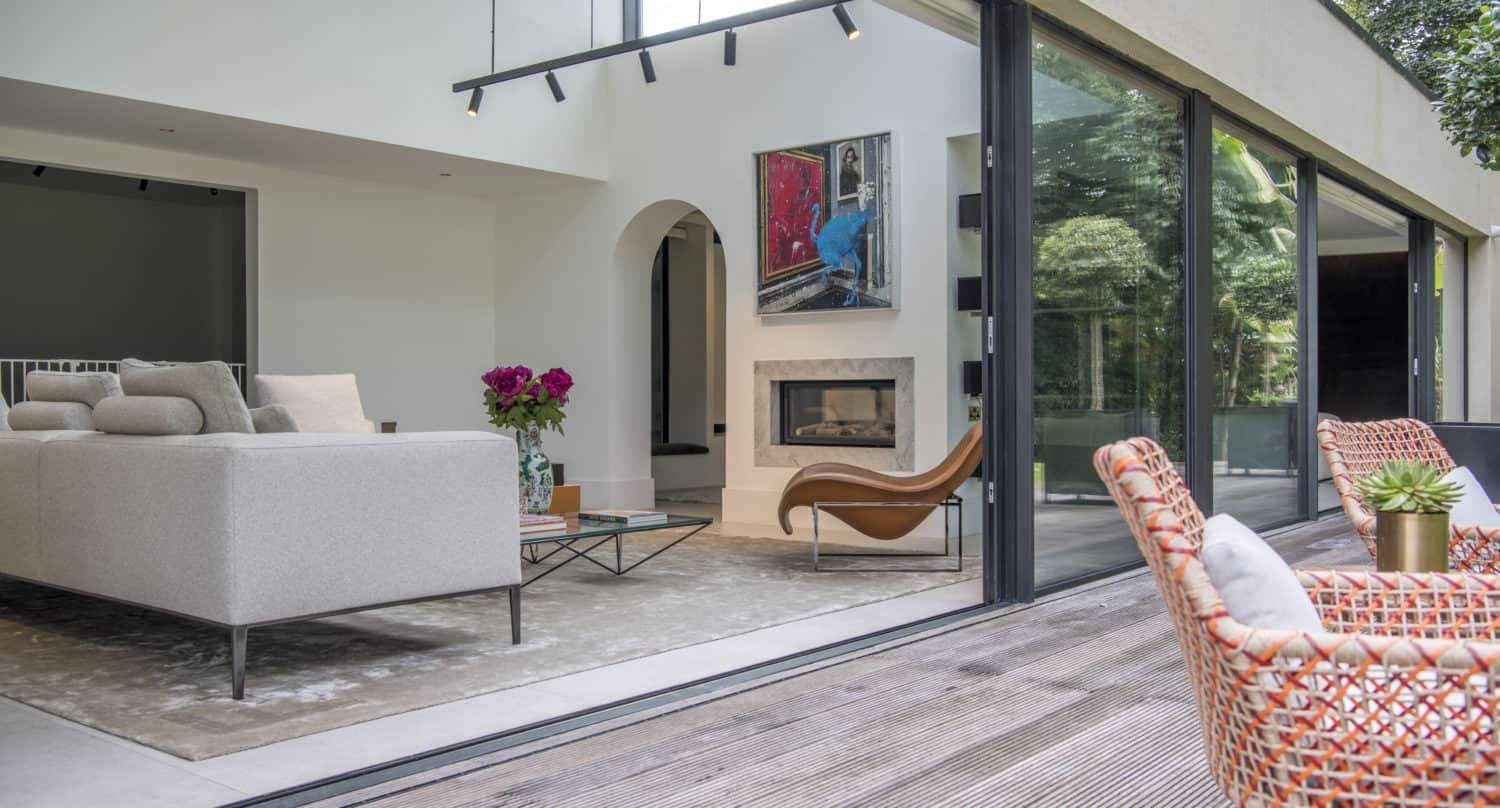 A modern living room featuring bespoke glass solutions with large sliding doors opening to a wooden deck. Inside, a light gray sofa, fireplace, artwork, curved chair, and flowers; outside, two orange woven chairs among lush plants.