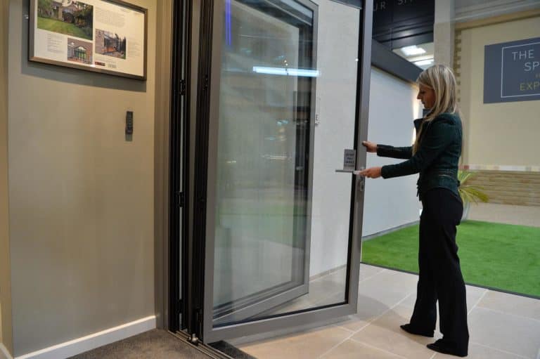 A woman with long blonde hair, wearing dark clothing, stands indoors holding open a large folding glass front door. The setting appears to be a modern showroom with green artificial grass outside.