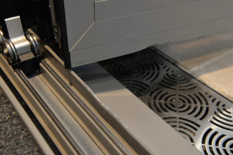 Close-up of a sliding door track system, showing the edge of bifold doors, metal rollers, the rail, and a decorative black metal drainage grate set into the floor beneath the door.