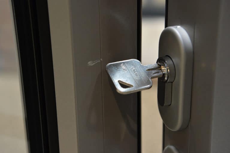A close-up view of a silver key inserted into the lock of a grey metal front door, with the edge of the door slightly open. The background is blurred.