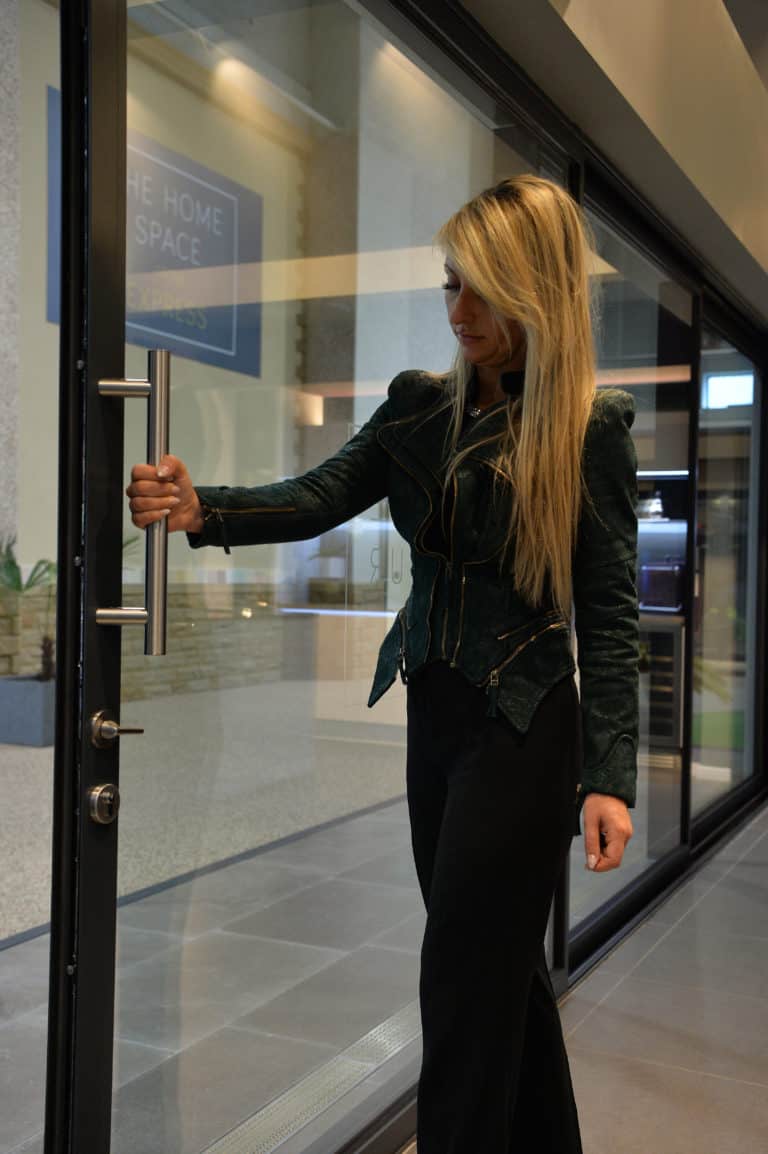 A woman with long blonde hair, wearing a dark jacket and black pants, is opening sleek sliding doors inside a modern building with large windows and a tiled floor.