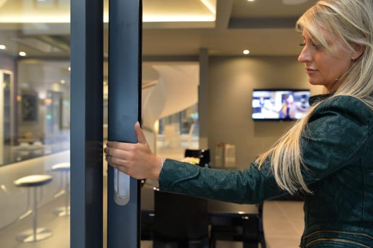 A woman with long blonde hair and a green jacket opens a bespoke glass solution front door in a modern, well-lit office or showroom, with a TV screen and seating visible in the background.