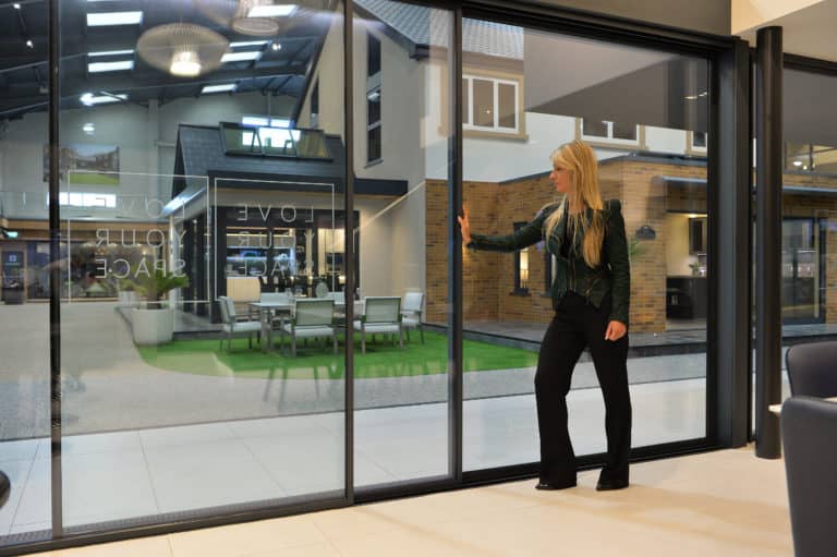 A woman with long blonde hair stands indoors, touching sleek sliding doors. Behind the glass, a modern outdoor seating area and a brick building facade highlight bespoke glass solutions in contemporary design.