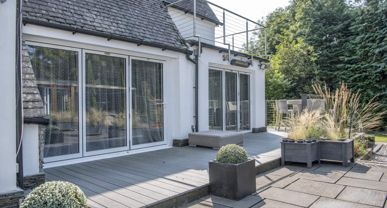 Modern house with large glass sliding doors and glass roofing opening onto a spacious gray deck, outdoor seating area, and several square planters with decorative grasses and shrubs. Lush greenery surrounds the patio.