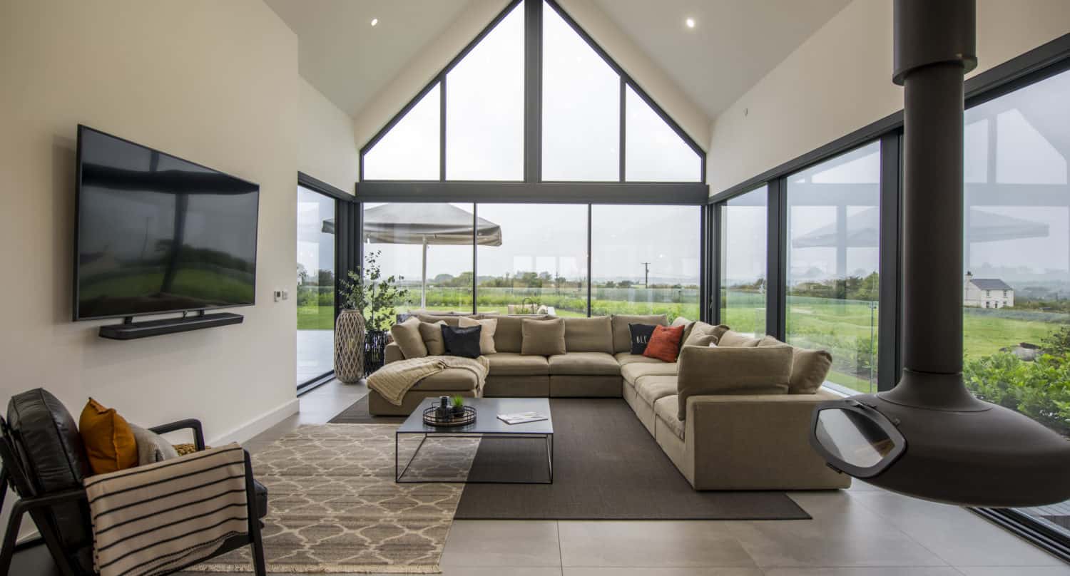 Modern living room with a beige sectional sofa, large windows offering scenic countryside views, a wall-mounted TV, contemporary fireplace, bifold doors, and minimalistic decor. Natural light fills the spacious, open area.
