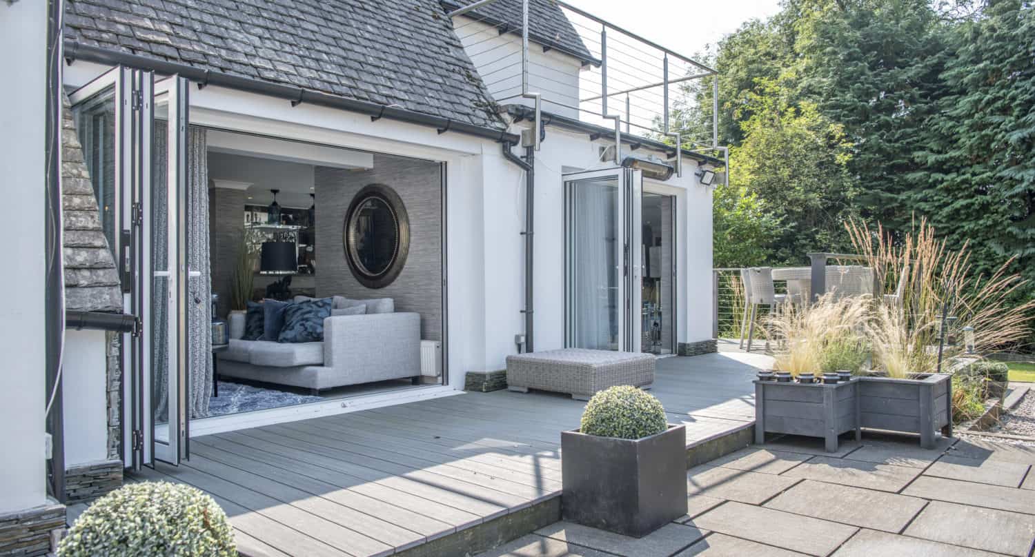 Modern house with bespoke glass solutions features large glass doors opening to a spacious patio, revealing a light-filled living room with a gray sofa. Potted plants decorate the patio, and trees and greenery surround the outdoor area.