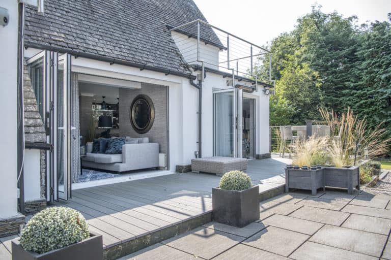 Modern house with bespoke glass solutions features large glass doors opening to a spacious patio, revealing a light-filled living room with a gray sofa. Potted plants decorate the patio, and trees and greenery surround the outdoor area.