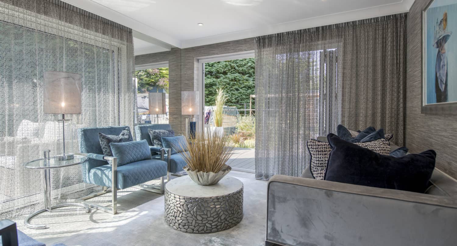 Modern living room with gray walls, blue armchairs, a round textured coffee table with tall dried grass centerpiece, sheer curtains, two lamps, and large windows with sliding doors overlooking a green garden. A painting hangs on the right wall.